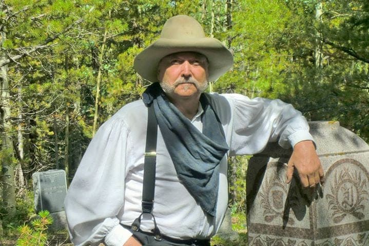 A reenactor in a cowboy hat, bandana and suspenders leaning on an ornate granite headstone