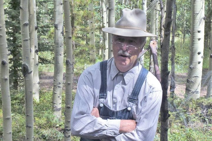 An older man in a fedora, round glasses and overalls portraying a miner in an aspen grove