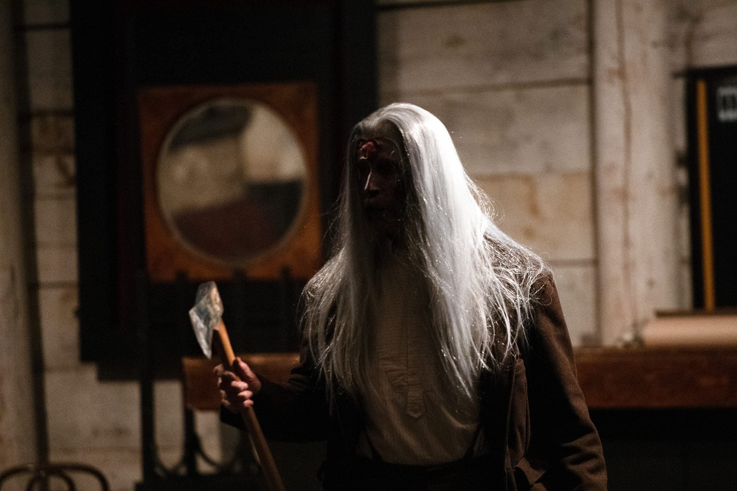 A Creepy Crawl guide with long white hair and a bloodied brow holds an axe in a dim parlor