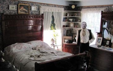 A Victorian bedroom with a tall carved bed, patterned wallpaper and a mannequin in a vest