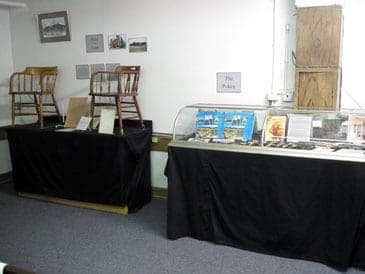 An exhibit room with antique chairs on draped tables, a case of books and a sign reading The Police