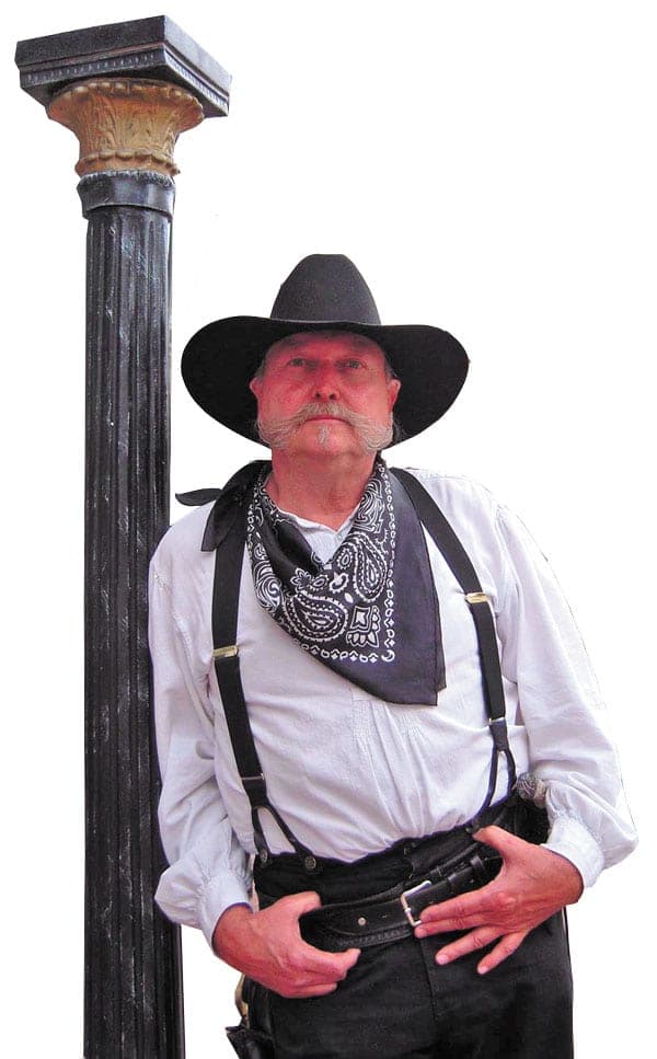 A mustached gunfighter in a black hat, paisley bandana and suspenders leaning on a cast-iron column