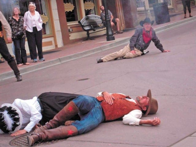 A staged shootout: two gunfighters lie in the street while spectators watch